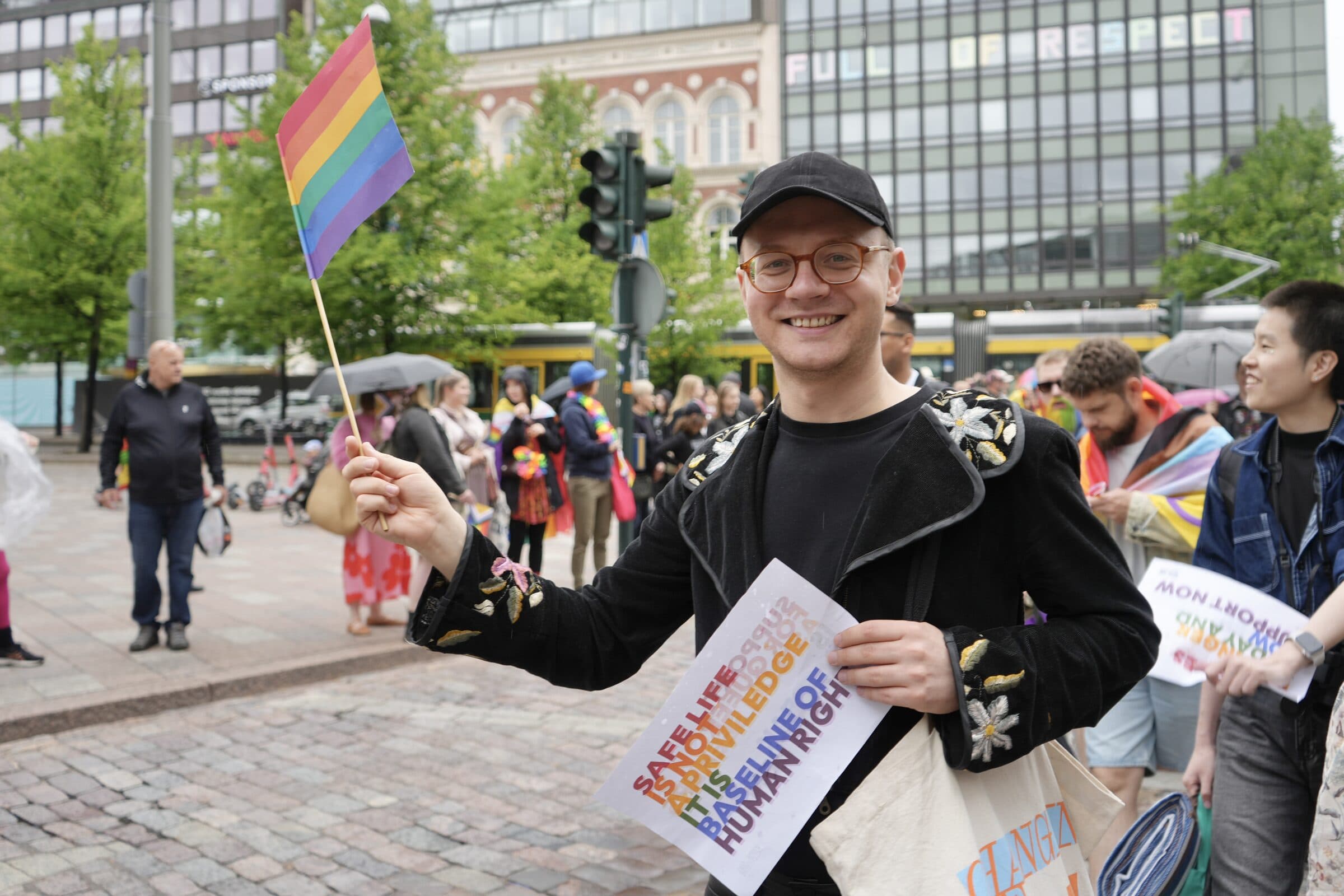 Hymyilevä henkilö pitää sateenkaarilippua ja kylttiä Helsingin keskustassa.