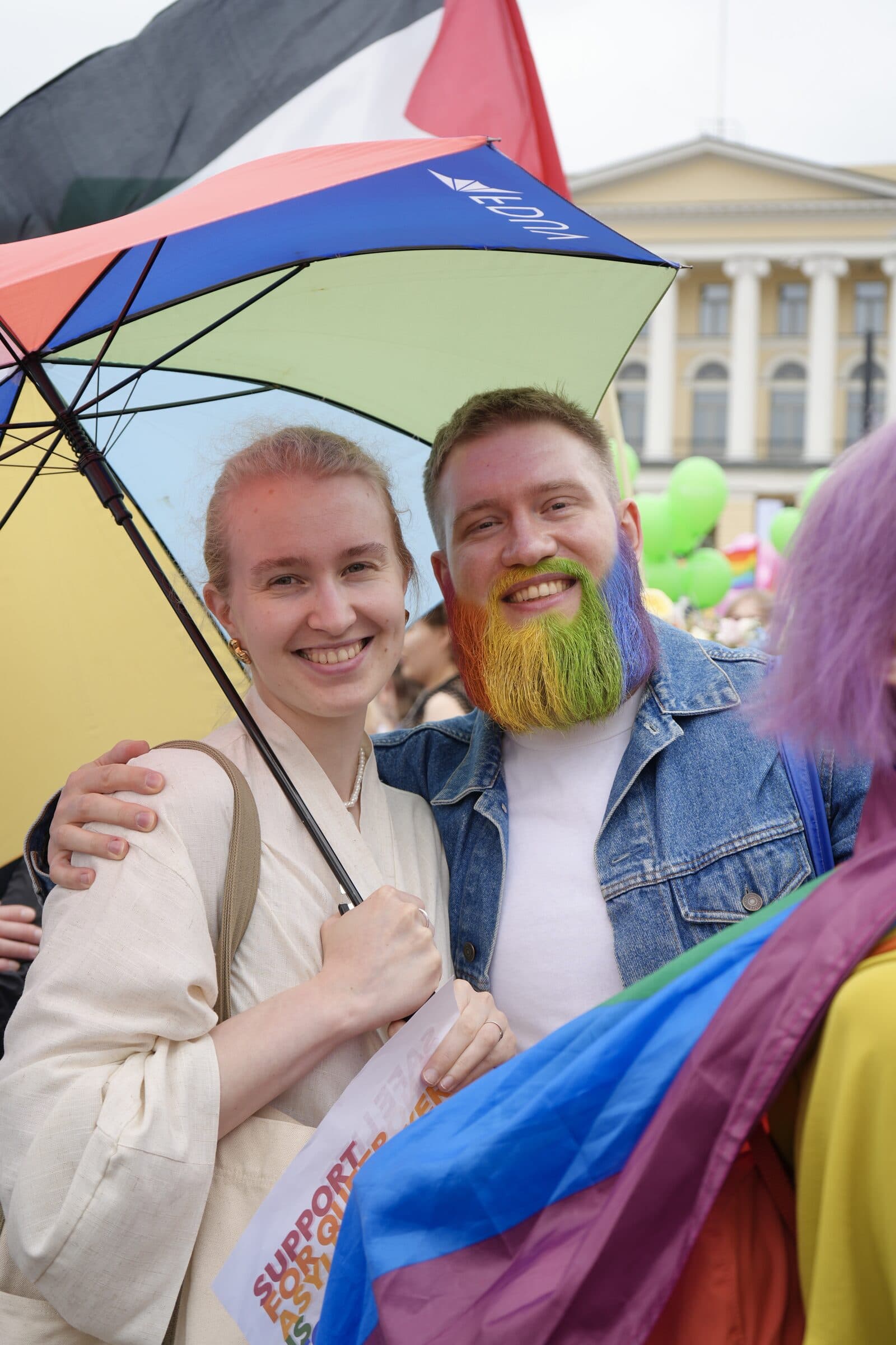 Kaksi hymyilevää ihmistä sateenvarjon alla Pride-päivänä, toisella sateenkaaren värinen parta.
